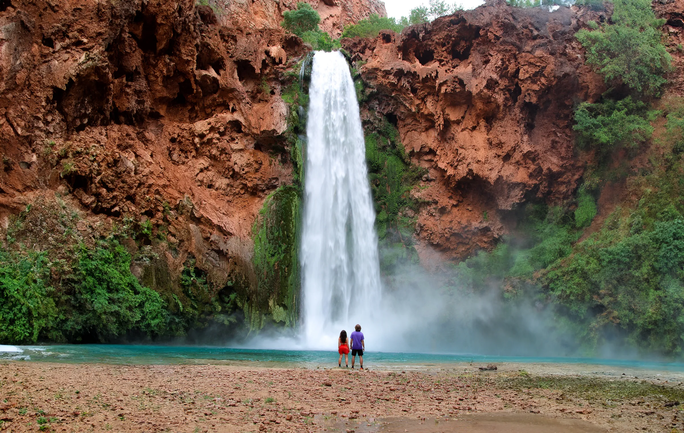 Havasu Falls pouring over red cliffs into a turquoise pool, two people on the shore.